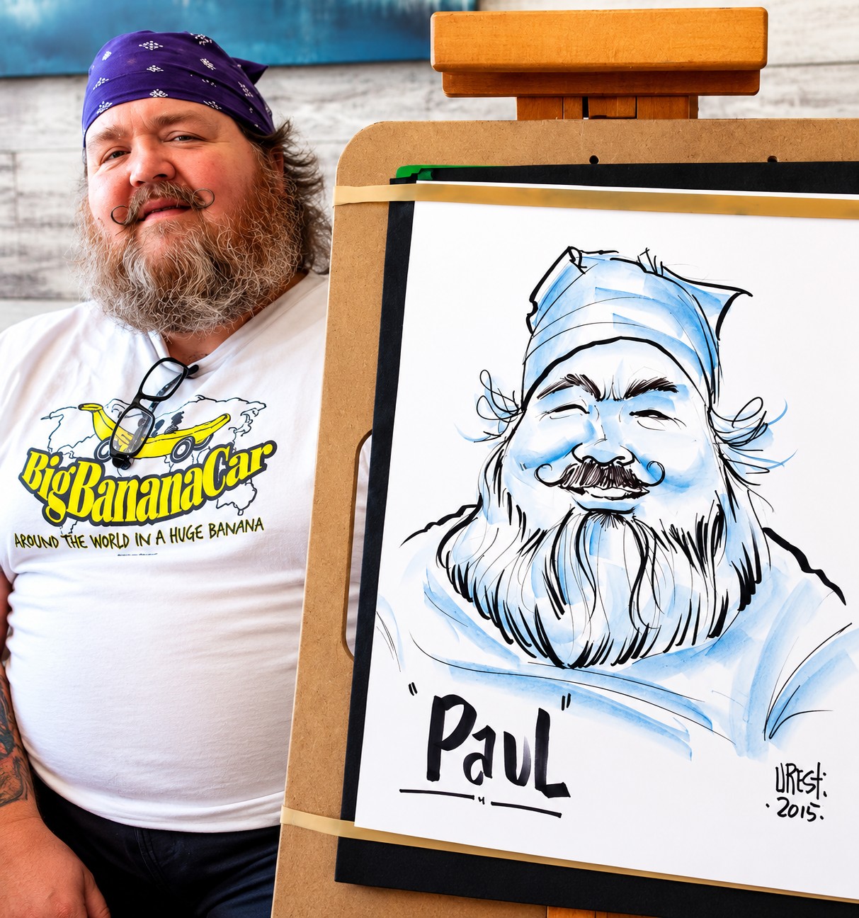 Eddie posing next to an easel displaying a blue-toned caricature titled Paul