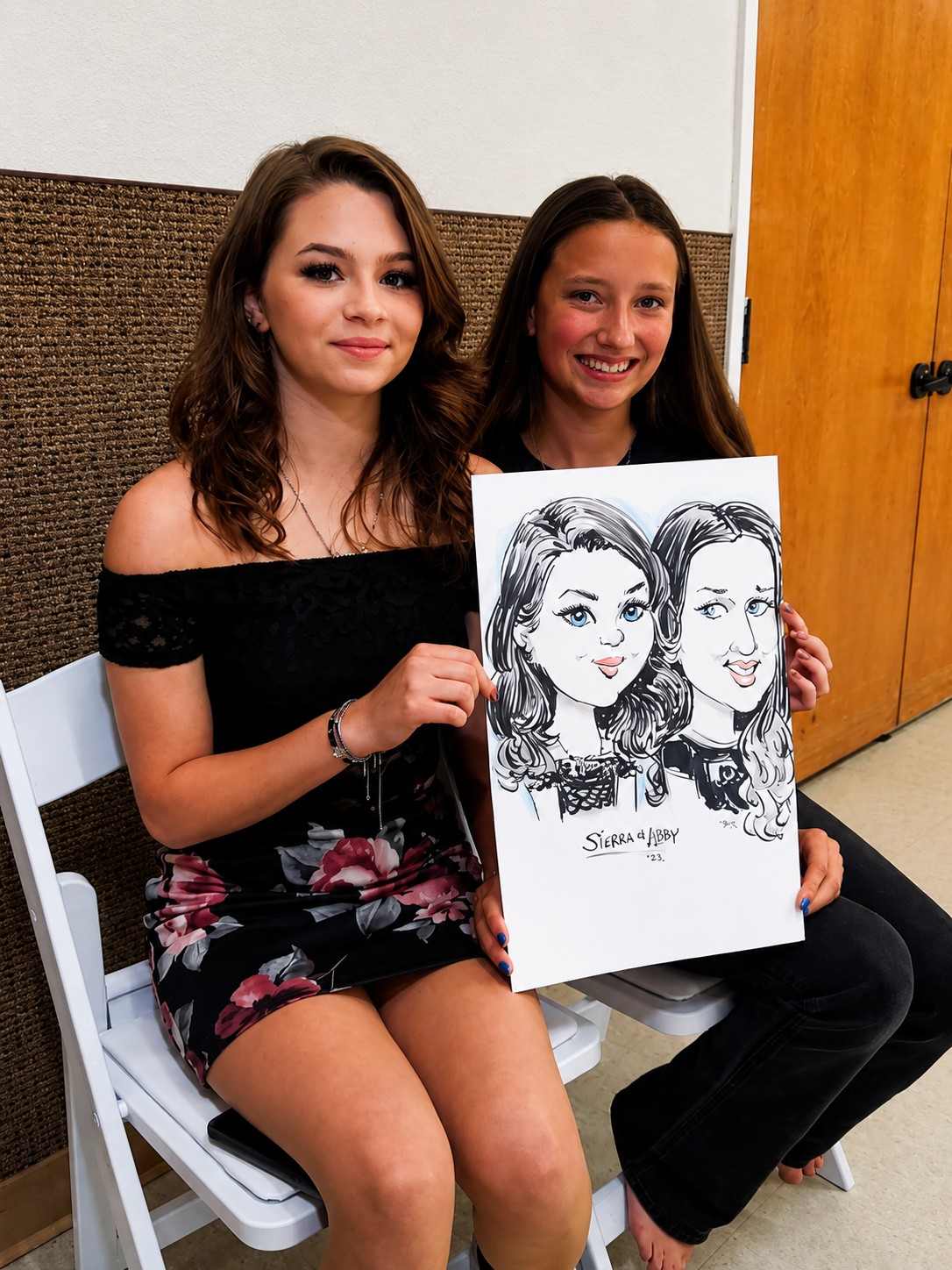 Two young women seated holding a duo caricature at a neighborhood get-together