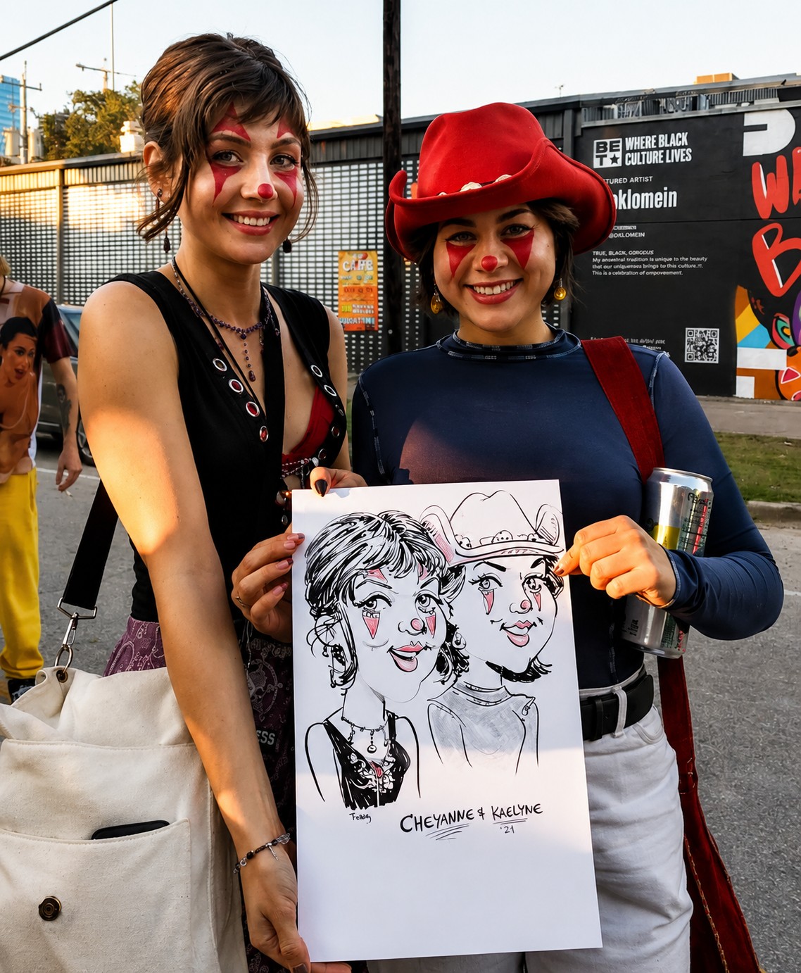 Two festival-goers in face paint holding a duo caricature at an outdoor comedy festival