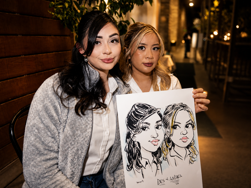Two women holding a duo caricature at an evening client appreciation event