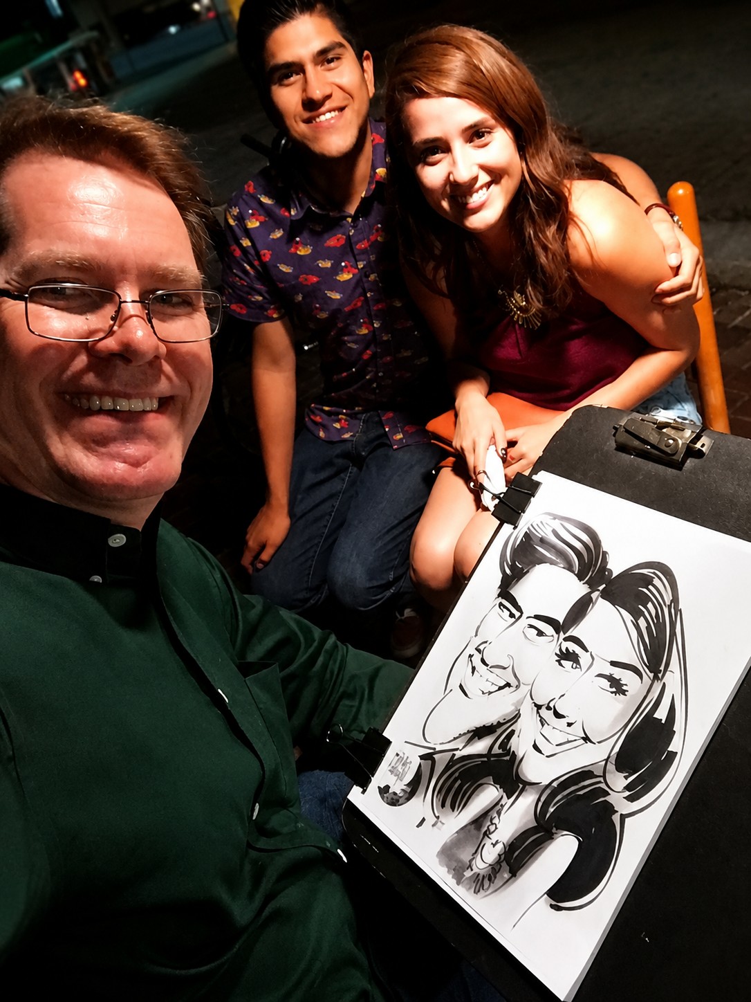 Jody with a couple holding a black-and-white duo caricature at an evening event